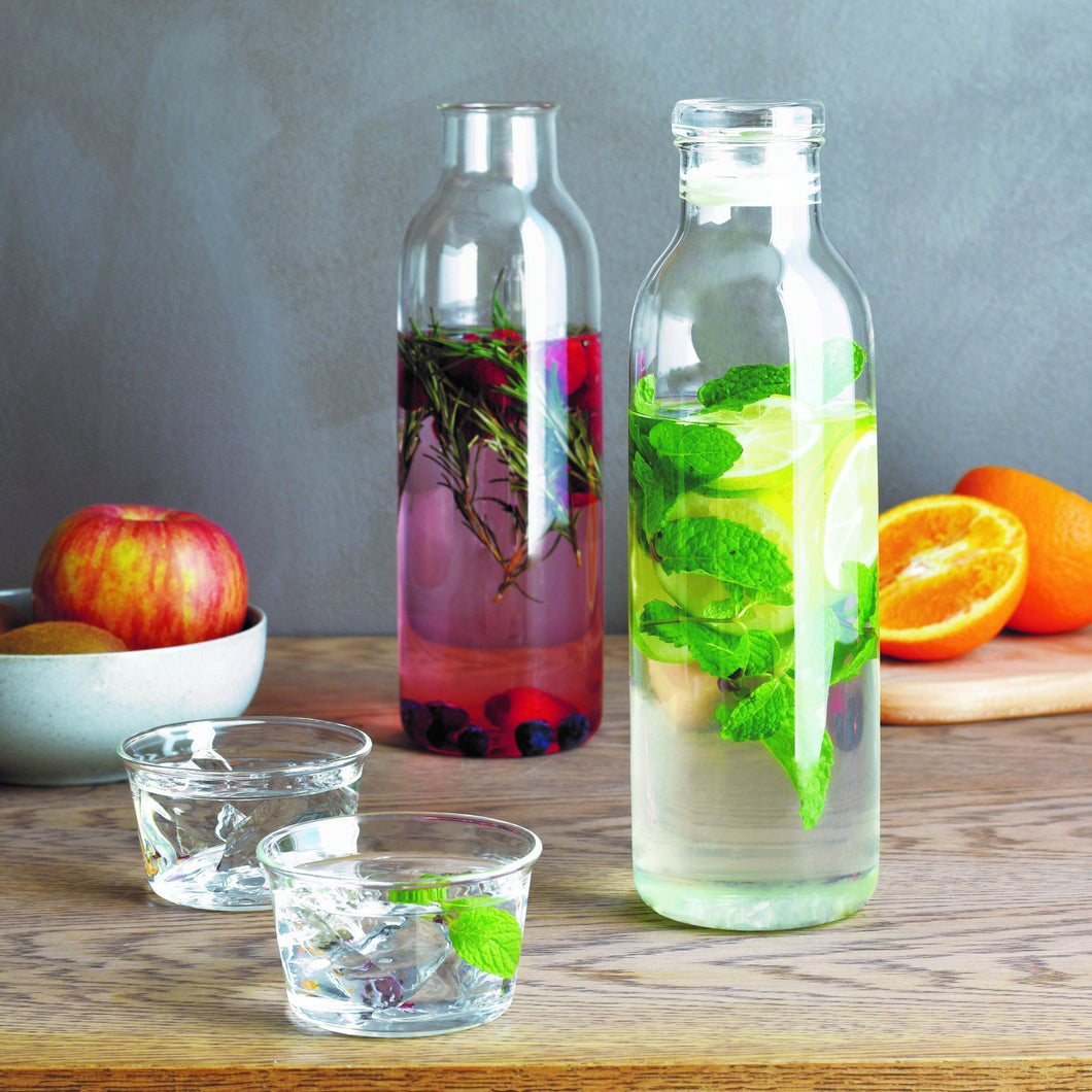 two kinto bottlit carafes with different flavored water inside them placed on a table with fruit and cups